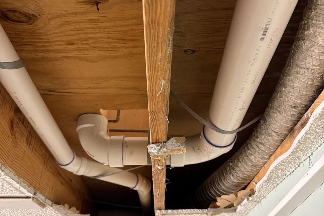PVC drain lines under floor joists in an opened ceiling beside a flexible duct