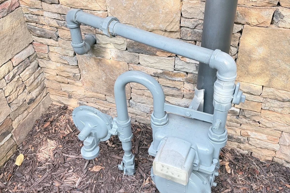 Gray painted gas meter, regulator, and pipes against a stacked stone wall over mulch