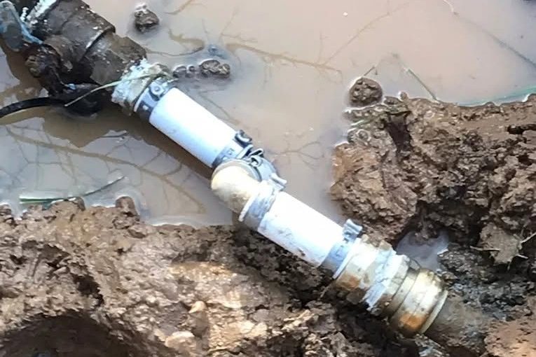 Muddy water-filled hole with a black pipe, white PVC, and a brass coupling joined for a water line repair