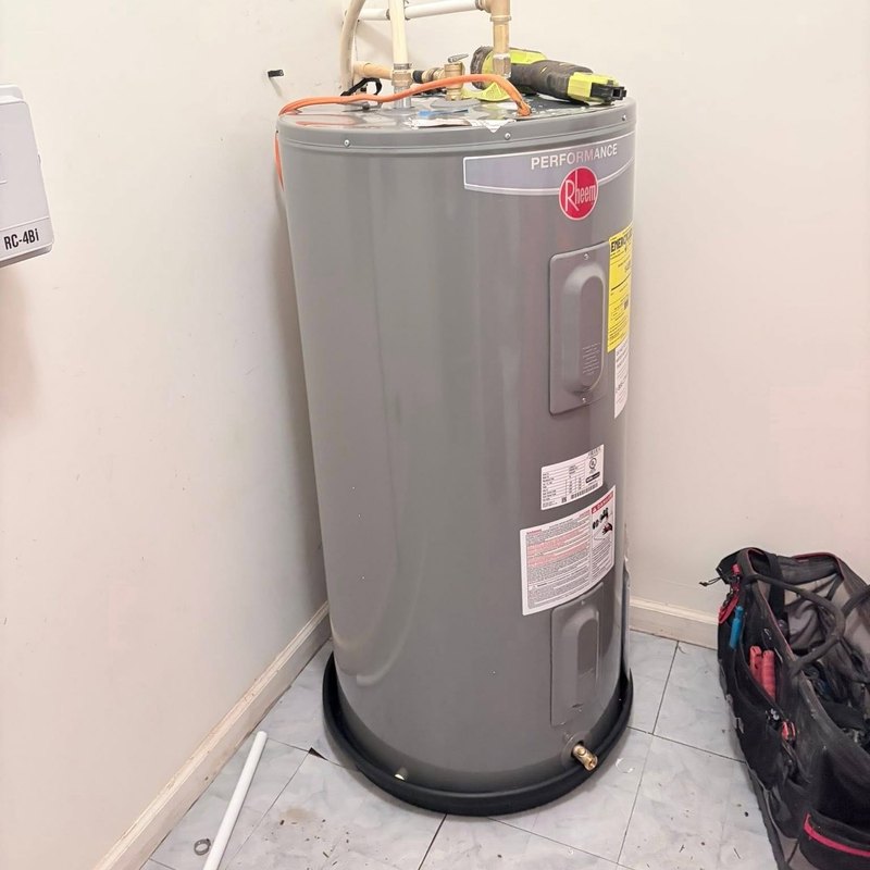 New gray electric tank water heater in a bathroom corner with a black tool bag beside it