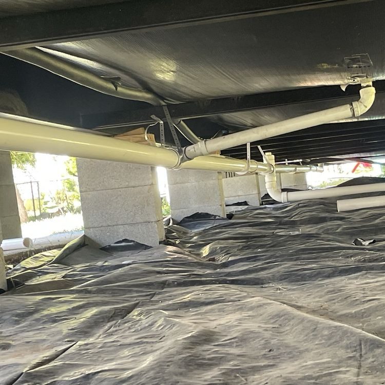 White PVC drain lines hanging beneath a floor over a black vapor barrier