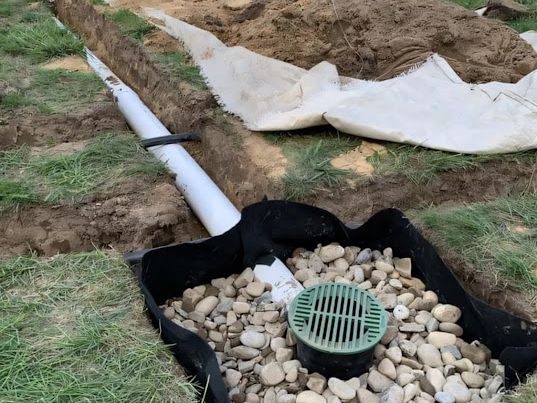Yard drainage trench with white PVC pipe leading to a green-grated catch basin in river rock