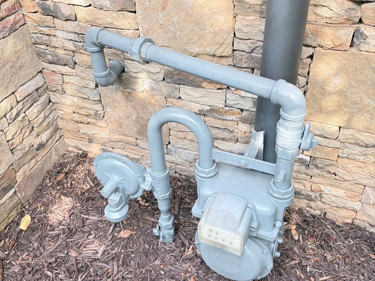 Gray painted gas meter, regulator, and pipes against a stacked stone wall over mulch
