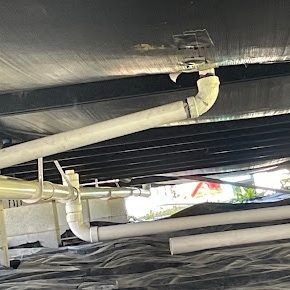 White PVC and iron drain lines hanging in a crawlspace under black vapor barrier