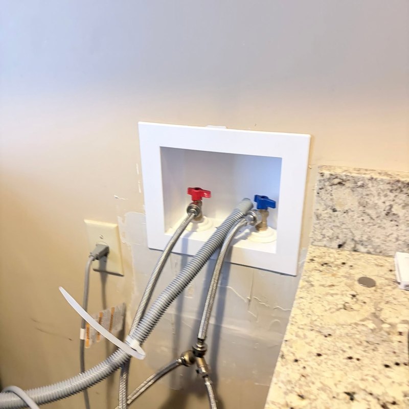 White washing machine outlet box with red and blue valves and braided hoses beside a granite countertop