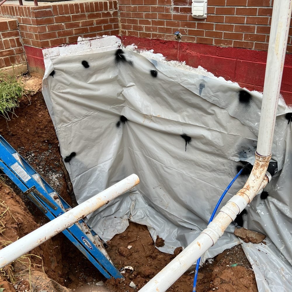 Excavation along a red-painted block wall with white PVC sewer pipe and a blue water line, a blue ladder in the hole