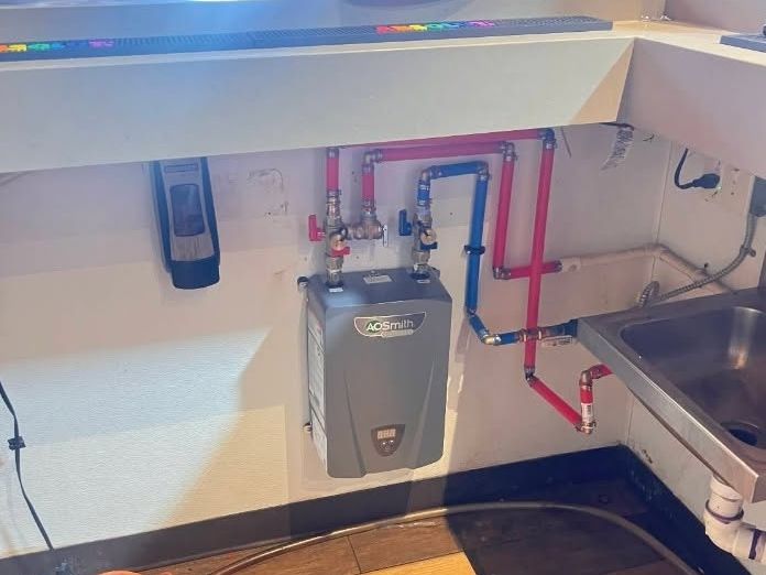 Small electric water heater mounted under a counter with red and blue PEX and a hand sink