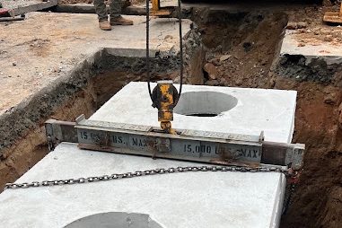 Concrete tank being lowered by chain into a gravel-lined excavation beside a truck