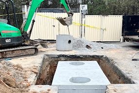 Two-chamber concrete tank set in a commercial lot excavation with a green mini excavator behind it