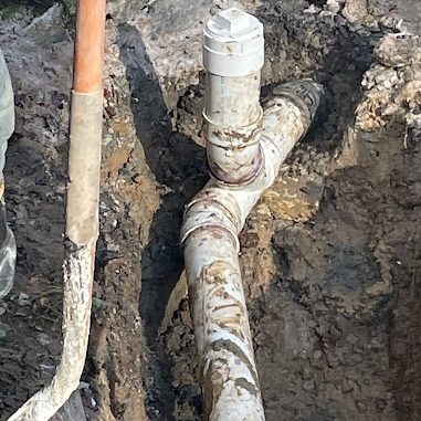 Muddy excavation showing a PVC cleanout and wye fitting joined to a sewer line, with a shovel handle in the foreground