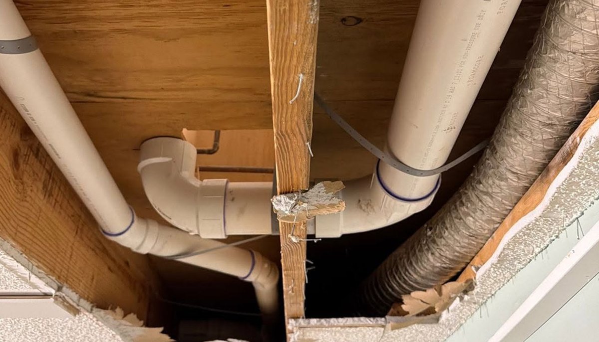 PVC drain pipes and a trap under floor joists in an opened ceiling with a duct beside them