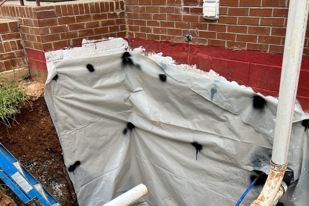 Excavation along a red-painted block wall with white PVC sewer pipe and a blue water line, a blue ladder in the hole