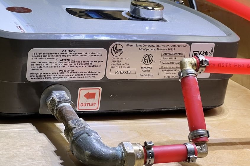 Compact electric water heater on a wall with red PEX piping and a rating label