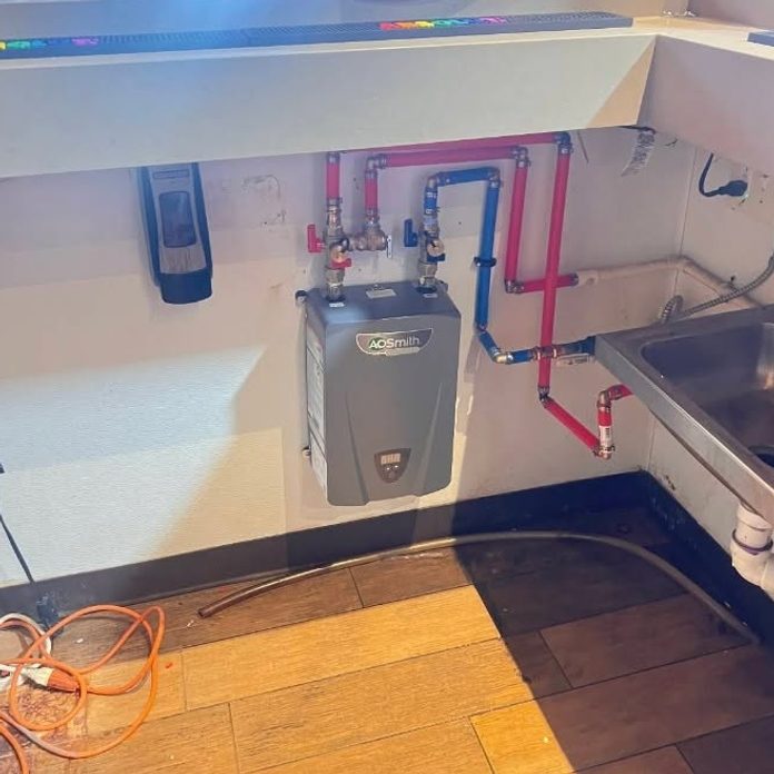 Small electric water heater mounted under a counter with red and blue PEX and a hand sink