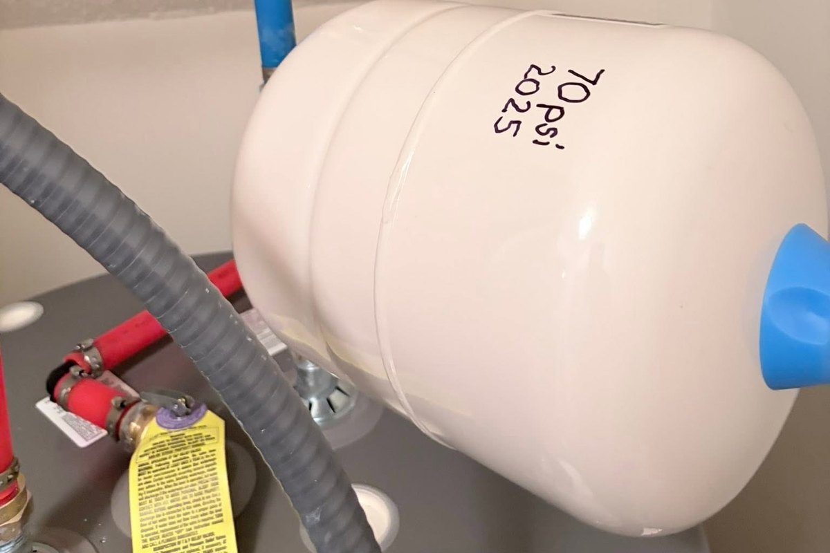 White thermal expansion tank marked 70 PSI installed above a water heater with blue PEX and a brass ball valve