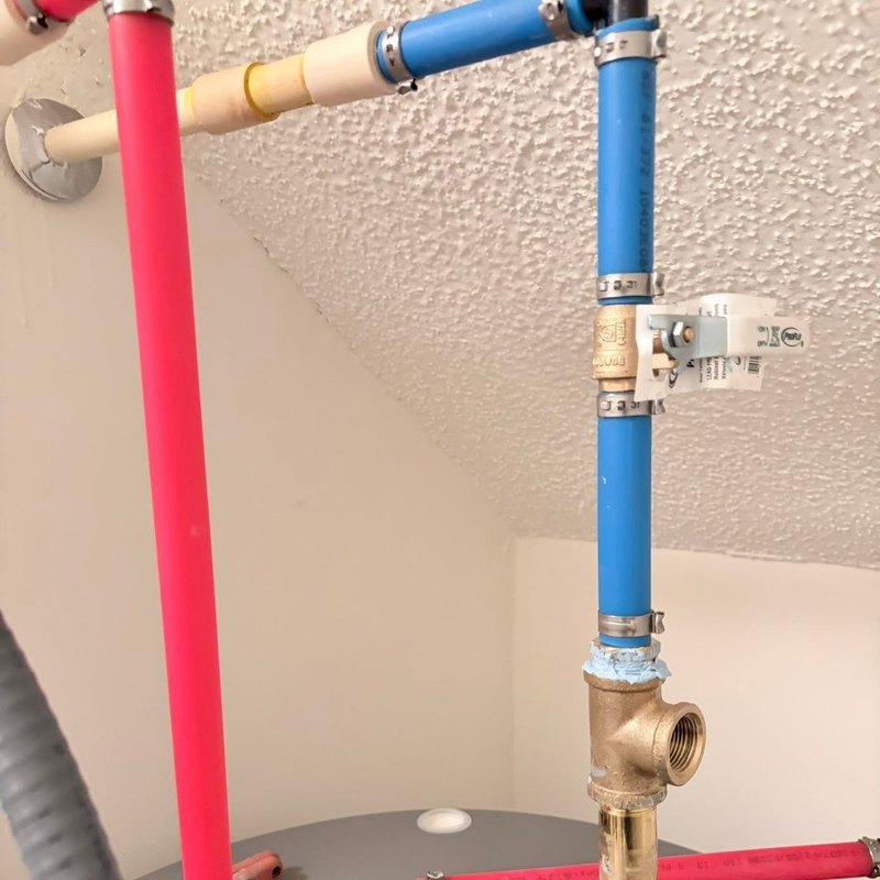 Closeup of red and blue PEX lines, a brass ball valve, and pressure relief piping on top of a gray tank water heater